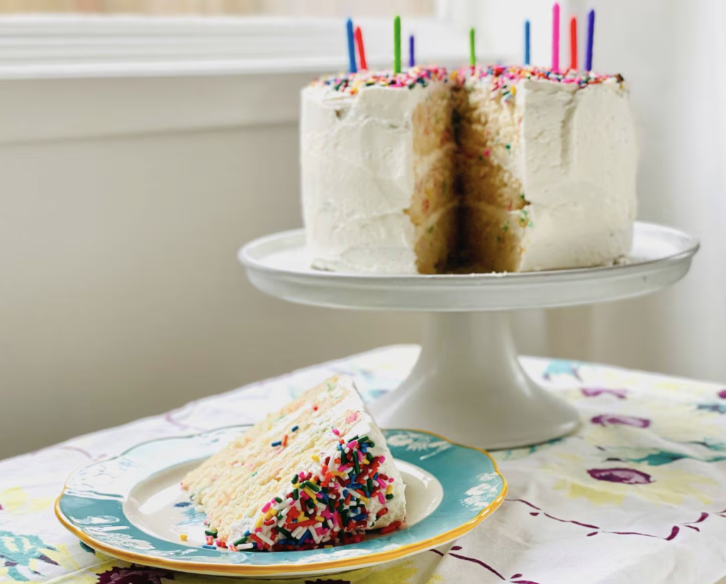 A slice of cake, with the top covered in sprinkles on a plate, with a large cake with candles in it sitting on a cake plate in the background